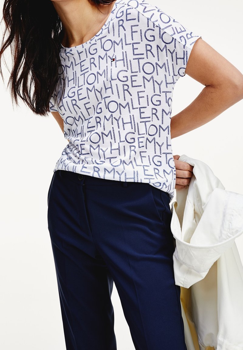 Camisa blanca con texto navy en un patrón repetido, combinada con pantalones azul oscuro. Una chaqueta ligera está colgada de un brazo.