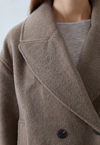Brun uldfrakke med et tekstureret stof, der har en spidse krave og to sorte knapper foran. Den indre lag er grå.