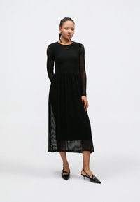 Black long-sleeve dress featuring textured, sheer fabric with horizontal stripes and a flared skirt, paired with black pointed-toe heels.
