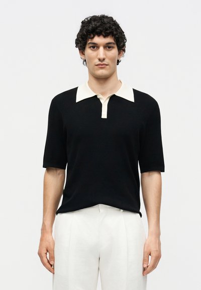 Homme aux cheveux bouclés portant un polo noir à manches courtes avec un col blanc et un pantalon blanc, debout devant un fond uni.
