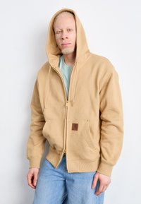 Beige zip-up hoodie made of thick cotton with a front pocket, gold zipper accents, and a logo patch. Worn with light blue jeans.