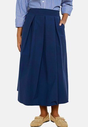 Žena oblečená v námornícky modrej midi sukni s plisovaním a vreckami, v kombinácii s modro-bielou pruhovanou košeľou a béžovými mokasínami.