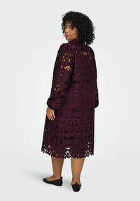 Čipkasta haljina u dubokoj ljubičastoj boji s složenim cvjetnim izrezima, nabranim rukavima i valovitim rubom. Nosena s crnim balerinkama, prikazana s leđa.