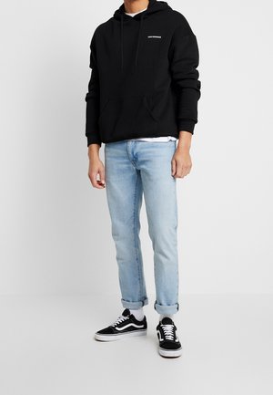 Zwarte hoodie met een voorzak, losse pasvorm en wit logo. Lichtblauwe jeans met opgerolde pijpen, gedragen met zwart-witte sneakers.
