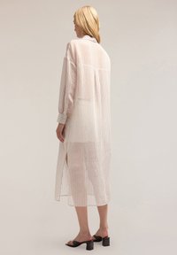 Abito lungo leggero e traslucido a strisce sottili in un morbido beige con linee verticali, maniche con bottoni e spacchi laterali, caratterizzato da una vestibilità rilassata.