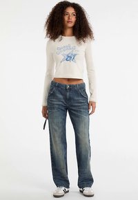 Top corto bianco a maniche lunghe con design grafico blu, abbinato a jeans in denim blu a lavaggio medio con taglio dritto e tasche.