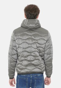 Giacca imbottita grigia con design trapuntato, cappuccio e polsini elasticizzati. Realizzata in materiale lucido e liscio. Indossata sopra jeans blu, vista da dietro.