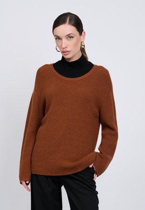 Donna con maglione marrone lavorato a maglia sopra un dolcevita nero e pantaloni neri, indossa grandi orecchini a cerchio dorati, in piedi davanti a uno sfondo chiaro e uniforme.