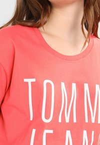 Korálové bavlněné tričko s kulatým výstřihem a bílým potiskem s nápisem "TOMMY JEANS" v tučném, ležérním písmu. Hladká textura a volný střih.