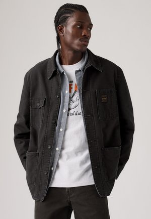 Uomo con capelli intrecciati che indossa una giacca nera sopra una camicia a righe con bottoni e una t-shirt bianca con grafica, guarda a sinistra con le mani nelle tasche.