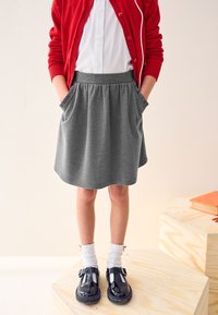 Gonna grigia con vita elasticizzata e tasche, indossata con una camicia bianca, un cardigan rosso, calze bianche e scarpe in vernice nera.