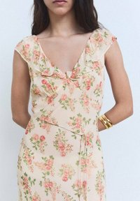 Robe fleurie en pêche pâle avec un col à volants, ornée de motifs floraux roses et verts sur un tissu doux ; soulignée par un détail de taille nouée.