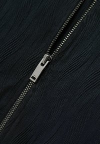 Tissu noir texturé avec un motif en vagues, arborant une fermeture éclair en métal argenté à l'avant, conçu pour une utilisation facile.