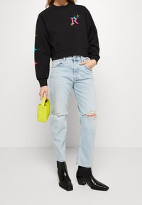 Felpa cropped nera con logo colorato, jeans distressed azzurri chiari, stivali neri alla caviglia e borsa piccola gialla brillante.