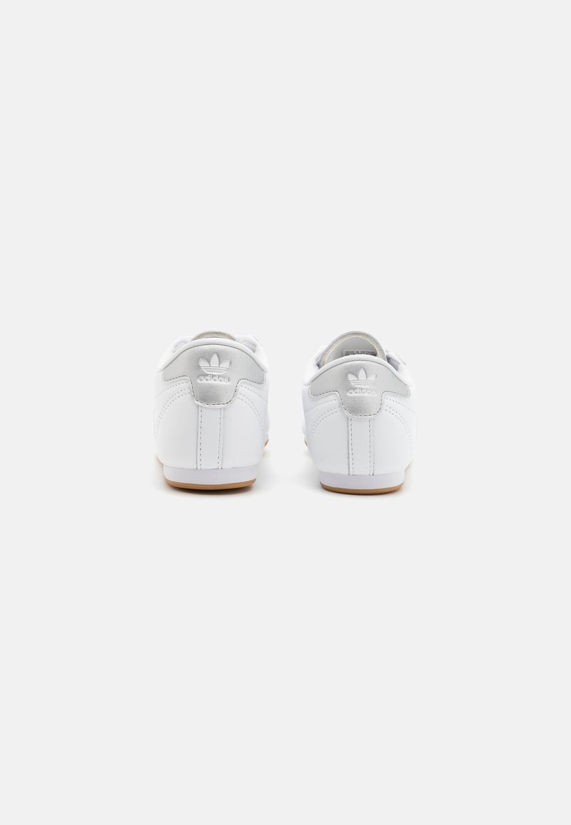 Białe sneakersy z gładką skórzaną cholewką, szarymi akcentami na pięcie i brązową gumową podeszwą. Posiadają klasyczne logo adidas z tyłu.