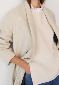 Manteau beige clair en mélange de laine avec manches larges et revers, superposé à un pull crème côtelé. Jean en denim bleu foncé visible.