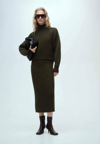 Gebreide olijfgroene tweedelige set met een cropped trui met hoge hals en een lange rok. Het model houdt een zwarte clutch vast en draagt donkere zonnebrillen.