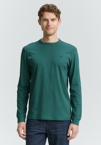 Langärmliges tealfarbenes Baumwoll-T-Shirt mit rundem Halsausschnitt, das ein kleines Logo auf der Brust trägt und einen lässigen Schnitt hat. Getragen mit dunklen Jeans.