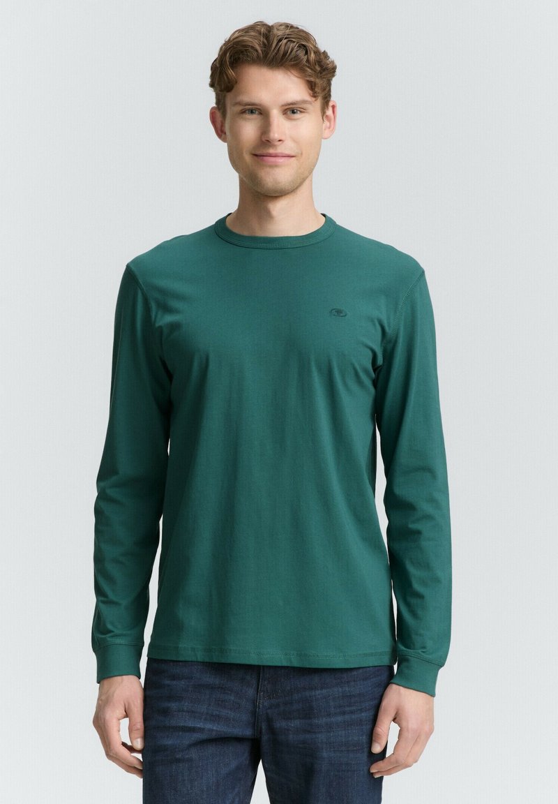Langärmliges tealfarbenes Baumwoll-T-Shirt mit rundem Halsausschnitt, das ein kleines Logo auf der Brust trägt und einen lässigen Schnitt hat. Getragen mit dunklen Jeans.