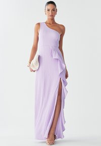 En axelband klänning i lavendel med texturerad tyg, en sidoflik och ett högt slits. Den kompletteras med en vit vävd clutch och beige sandaletter med remmar.