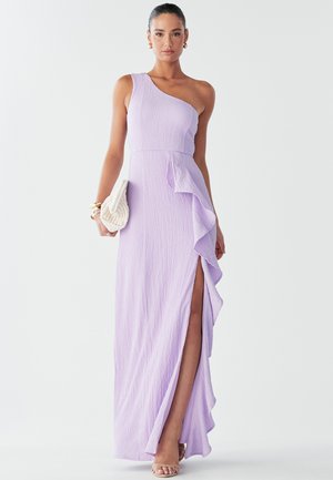 Vestido lila de un solo hombro con tejido texturizado, un volado lateral y una abertura alta en el muslo. Incluye un clutch blanco de trenzado y tacones beige con tiras.