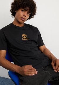 Schwarzes Baumwoll-T-Shirt mit Rundhalsausschnitt und kurzen Ärmeln, mit einem orangen Timberland-Logo-Graphic auf der Vorderseite.