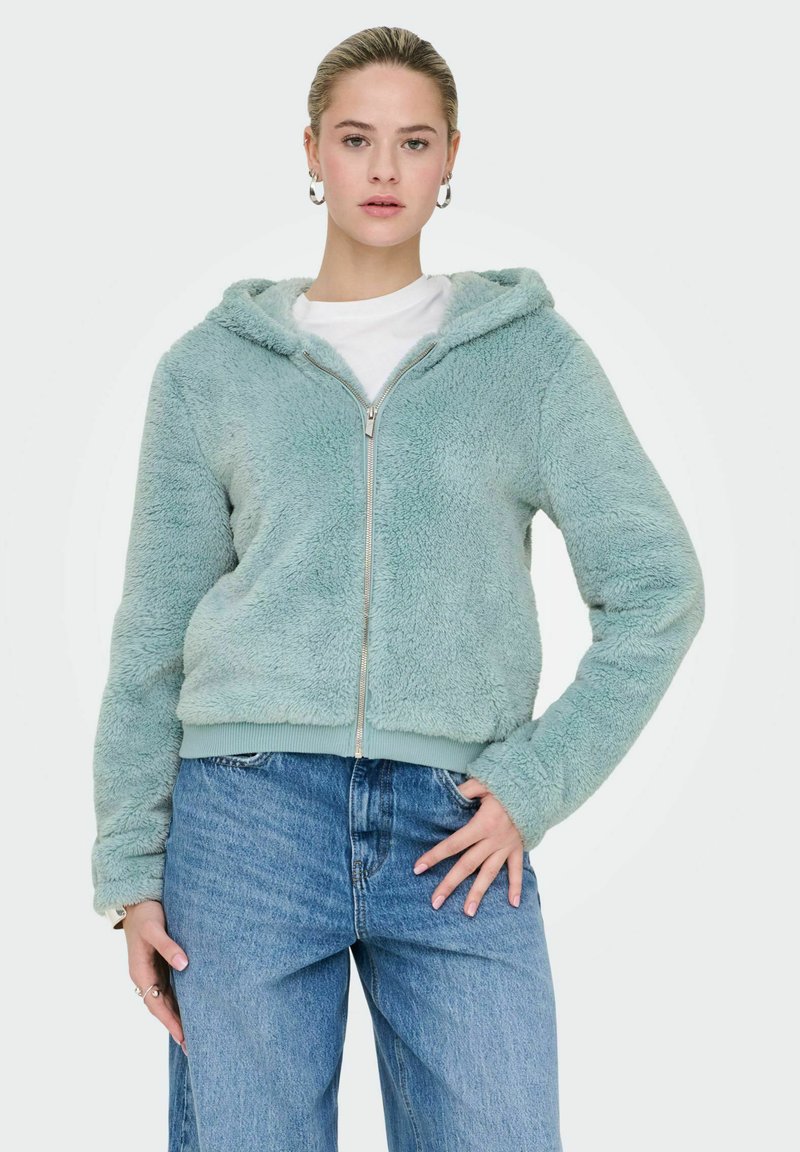 Faux fur zip-up hoodie in light mint green, featuring a lined hood, ribbed hem, and long sleeves. Paired with loose blue denim jeans.