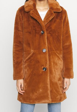 Manteau en fausse fourrure marron avec trois boutons noirs, col large, manches longues et poches latérales, porté sur un jean bleu clair.