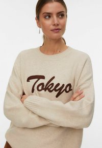 Vrouw met lichtbruin haar dat in een staart is vastgebonden, gekleed in een beige trui met de tekst "Tokyo", met gekruiste armen en kleine hangers in haar oren, tegen een witte achtergrond.