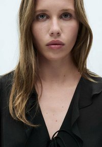 Black blouse featuring a ruffled neckline and a tie detail at the front, made from a textured fabric, worn by a person with long hair.