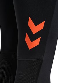 Czarne legginsy o gładkiej, rozciągliwej fakturze z pomarańczowym wzorem w kształcie litery V na udzie. Posiadają siatkowy wzór na dolnej części nogawki.