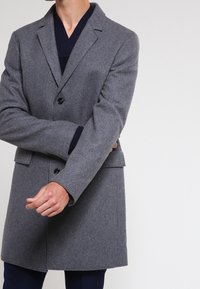 Abrigo de lana gris con corte ajustado, cierre de botón simple y bolsillos. La tela texturizada muestra un acabado suave y un diseño estructurado.