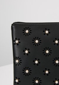 Petite pochette en cuir noir avec une fermeture éclair, décorée de motifs floraux découpés au laser et ornée de petites perles semblables à des perles.