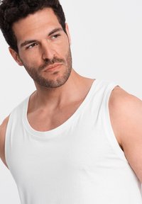 Débardeur en coton blanc avec un col rond et de larges emmanchures, présenté sur un modèle aux cheveux courts et foncés avec une légère barbe.