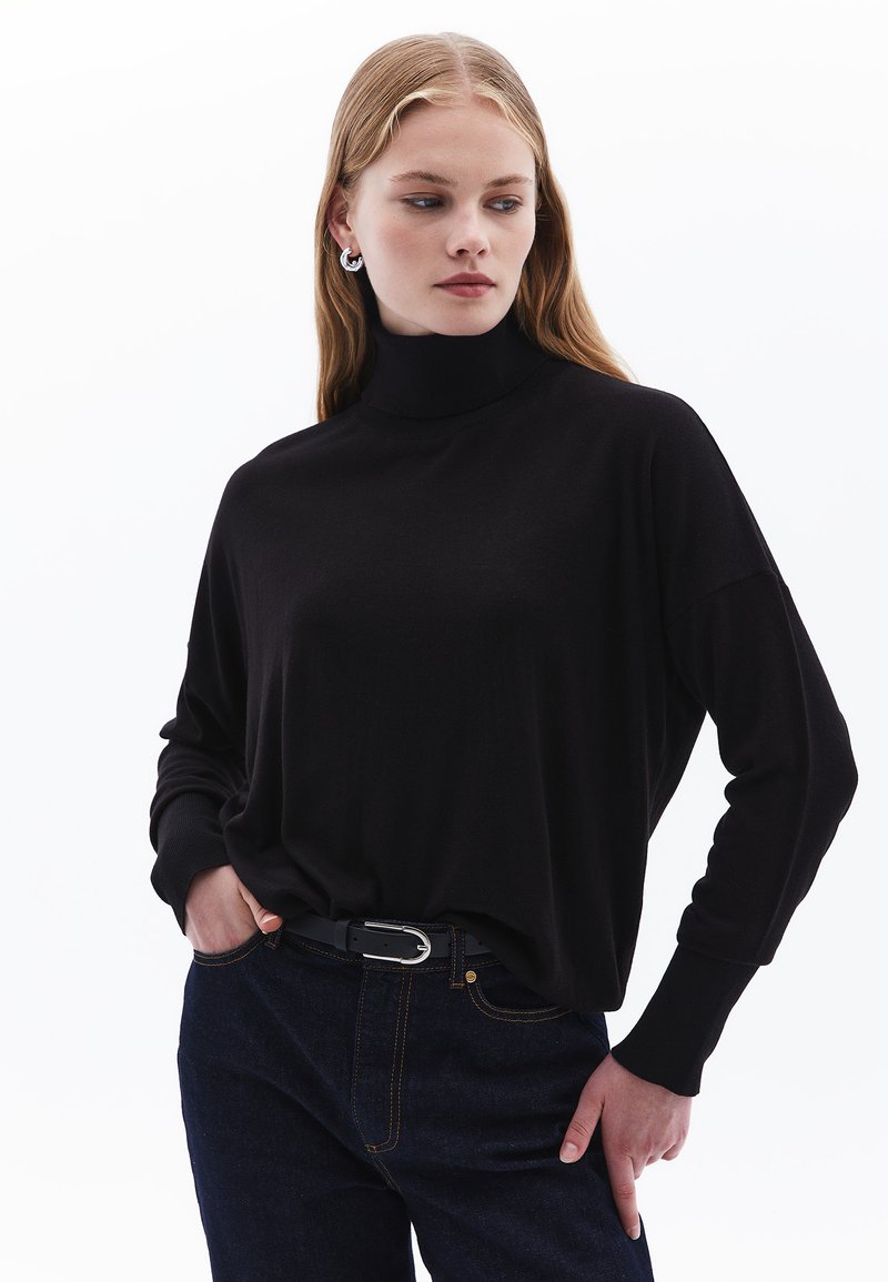 Schwarzer Rollkragenpullover aus weichem Stoff mit langen Ärmeln, lockerem Schnitt und gerippten Bündchen, kombiniert mit dunkelblauen Jeans und einem Gürtel mit silberner Schnalle.