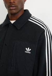 Černá fleece bunda s límečkem na knoflíky, třemi bílými pruhy na rukávech a vyšitým bílým logem Adidas na kapsičce na prsou.