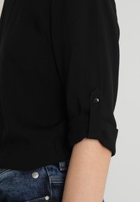 Person som bär en svart långärmad skjorta med uppvikt ärm som säkrats med knapprör och blå denimjeans.