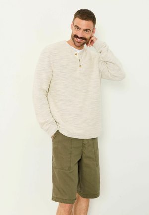 Lächelnder Mann mit Schnurrbart, der einen beige gestrickten Henley-Pullover und olivgrüne knielange Shorts trägt, steht mit einer Hand in der Tasche und geneigtem Kopf.