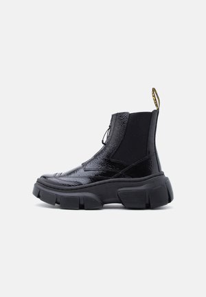 Bottine à plateforme épaisse noire brillante avec panneau élastique latéral et languette d'entraînement sur un fond blanc.