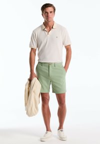 Korte mouwen crème polo met een contrasterende kraag, gecombineerd met lichtgroene shorts en witte sneakers. Een beige jack wordt in één hand vastgehouden.