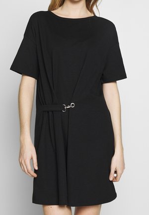 Femme portant une robe noire manches courtes avec un col rond et un détail de fermoir argenté cintrant la taille sur un fond uni.