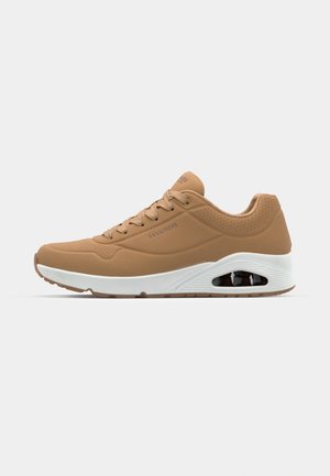 Tan sneaker with a smooth synthetic upper, perforated detailing, white midsole, and visible air cushioning in the heel. Flat laces.