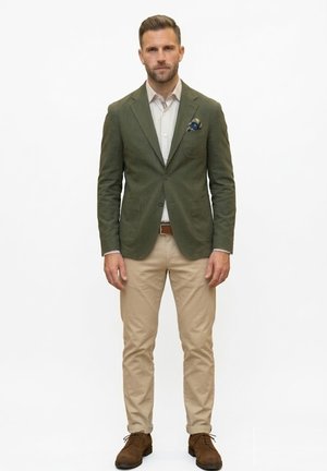 Hombre de pie, vestido con blazer verde oliva, camisa blanca con estampado, pantalones beige, cinturón marrón y zapatos de ante marrón con pañuelo en el bolsillo.