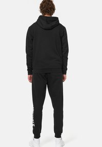 Zwarte hoodie en joggingbroek gemaakt van zachte stof. Hoodie met een grote achterkantzak, broek heeft zijzakken en een wit logo-details op het been.