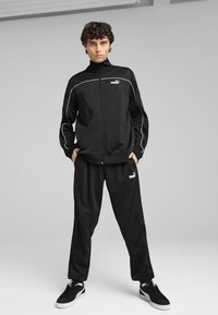 Chándal negro con chaqueta con cremallera y pantalones a juego. Presenta detalles en blanco y acentos de logo. La tela parece suave y ligera.