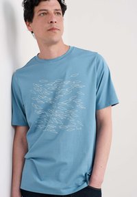 Camiseta de algodón azul claro con un patrón gráfico de peces blancos en la parte delantera. Cuello redondo y mangas cortas. Diseño de ajuste casual.