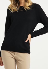 Schwarzer Strickpullover mit lockerer Passform, Rundhalsausschnitt und langen Ärmeln; kombiniert mit beigen Hosen. Hat einen gerippten Saum und seitliche Akzente.