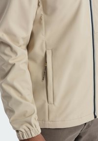 Beige lätt jacka med en mjuk textur, elastiska mudd, sidfickor och en svart dragkedja längs framsidan.