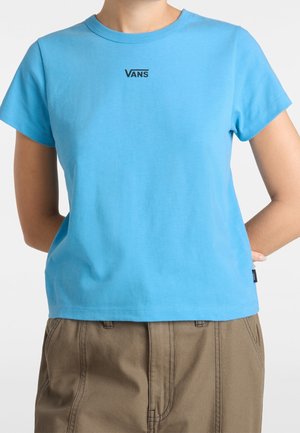 Osoba ubrana w jasnoniebieską koszulkę Vans z krótkim rękawem i brązowe spodnie, z rękami za plecami na białym tle.