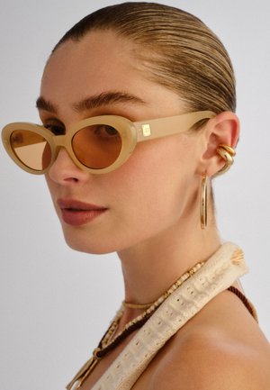 Femme aux cheveux tirés en arrière portant des lunettes ovales beiges, de grandes boucles d'oreilles en or, des colliers superposés de perles et un haut crème texturé.